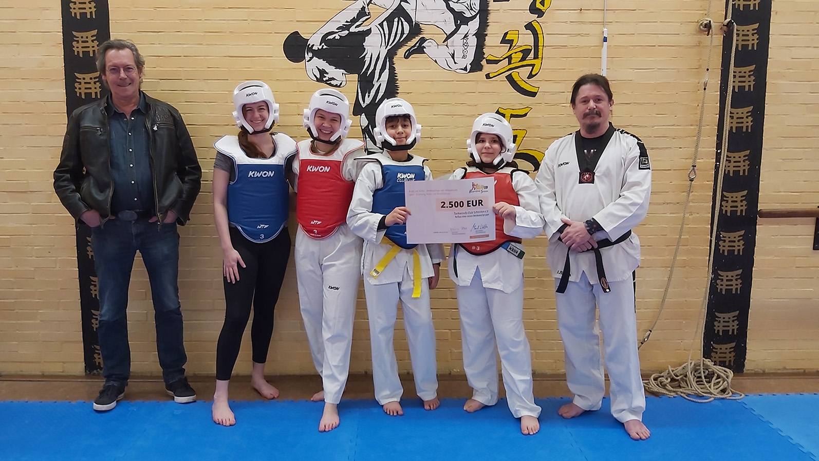 Stellv. Kuratoriumsvorsitzender Bernd Kehren (l.) überreichte gleich zwei Förderschecks der Bürgerstiftung Schleiden an den Taekwondo Club Schleiden e.V. die einige Sportler:innen sowie Trainer Mark Bauer (r.) entgegen nahmen und auch gleich die Wettkampfausrüstung präsentierten.