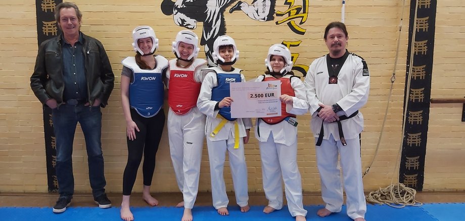 Stellv. Kuratoriumsvorsitzender Bernd Kehren (l.) überreichte gleich zwei Förderschecks der Bürgerstiftung Schleiden an den Taekwondo Club Schleiden e.V. die einige Sportler:innen sowie Trainer Mark Bauer (r.) entgegen nahmen und auch gleich die Wettkampfausrüstung präsentierten.