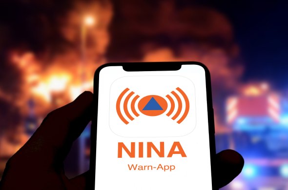 Smartphone zeigt NINA Warn-App, im Hintergrund die Szenerie eines Großbrands in der Nacht.